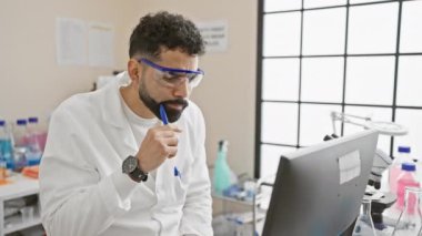 Laboratuar gözlüğü takan sakallı düşünceli bir adam laboratuvar ortamında bilgisayardaki verileri analiz ediyor..