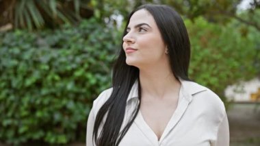 Uzun esmer saçlı, genç ve güzel bir İspanyol kadın yemyeşil bir parkta güven içinde poz veriyor..