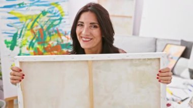 Gülümseyen olgun İspanyol kadın sanatçı renkli bir sanat stüdyosunun içinde boş bir tuval tutuyor..
