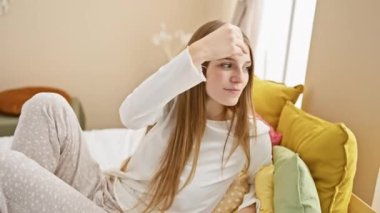 Çekici, kendine güvenen genç, sarışın bir kadın. Pijamalarıyla poz veriyor, yatağında oturuyor, şık bir şekilde saçlarına dokunuyor.