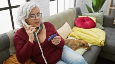 Yetişkin bir kadın, elinde kredi kartıyla telefonda konuşur. İçeride uyuyan bir köpeğin yanında bir kanepede oturur..