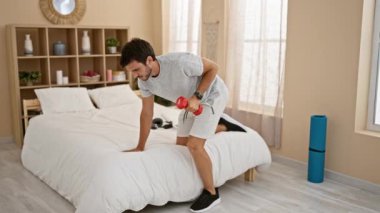 Dambıllarla egzersiz yapan odaklanmış bir adam düzenli bir yatak odasında.