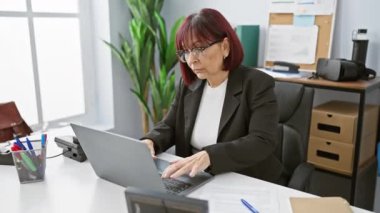 Orta yaşlı İspanyol bir kadın masasında, parlak bir ofis ortamında çalışıyor. Bilgisayarına, kırtasiyesine ve bitkilerine odaklanıyor..