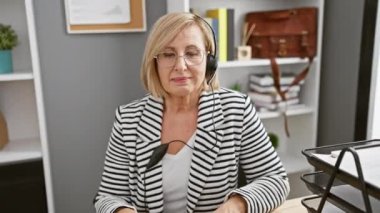 Gülümseyen, orta yaşlı, kulaklık takan, modern bir ofis odasında çalışan sarışın bir kadın..