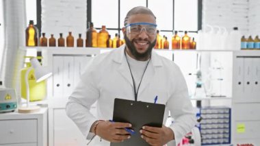 Sakallı, laboratuvar önlüğü ve koruyucu gözlük takan, gülümseyen Afro-Amerikan erkek bilim adamı..