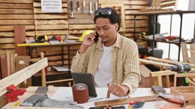 Genç adam ahşap atölyesinde telefon ve tabletle birden fazla iş yapıyor.