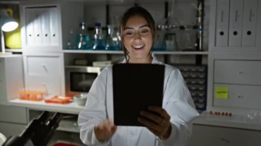 Laboratuvar önlüklü, gülümseyen İspanyol bir kadın laboratuvar ortamında bir tablet tutuyor, profesyonellik ve neşe simgeliyor..