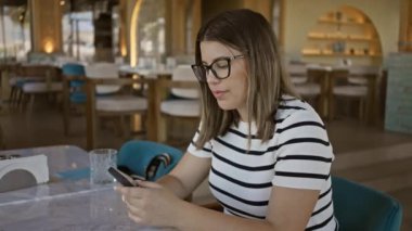 Parlak, modern bir restoranda akıllı telefon kullanan gözlüklü, gülümseyen genç bir kadın.