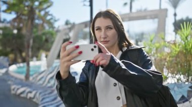 Beyaz kadın güneşli şehir caddesinde akıllı telefonuyla selfie çekiyor.