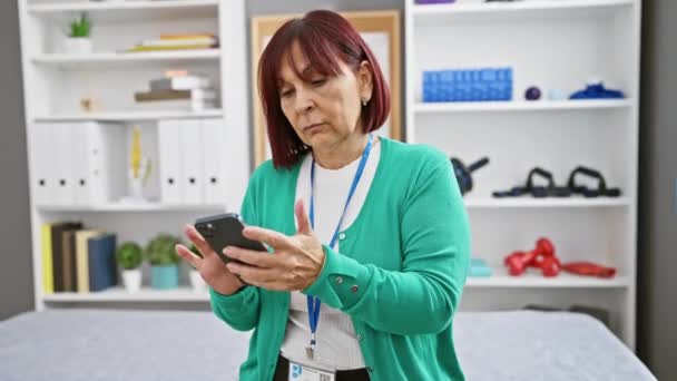 成熟した女性は 診療所のリハビリテーションルームでスマートフォンを使用し ヘルスケアの技術を示しています — ストック動画