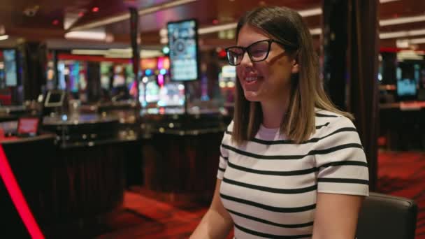 バックグラウンドで活気に満ちたスロットマシンでカジノで遊ぶ幸せな女性は レジャーと興奮を意味します — ストック動画