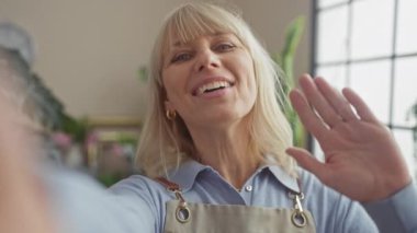 Çiçekçi önlüğündeki gülümseyen sarışın bir kadın evde selfie çekmekten hoşlanıyor.