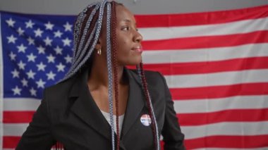 Kendine güvenen örgü örgülü, spor ceket giyen ve oy veren Amerikalı bir kadın bir ofisteki Amerikan bayrağının önünde duruyor..