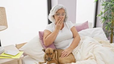 Şaşırmış gri saçlı orta yaşlı kadın köpekle yatakta oturuyor, ağzı ve elleri şokta. Pijama giyiyor, hata yapmaktan korkuyor. Yatak odasında şok edici bir ifade..