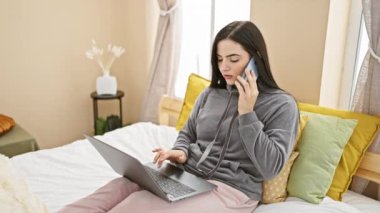 Genç bir İspanyol kadın rahat bir yatak odasında dizüstü bilgisayarla konuşurken telefonla konuşuyor..