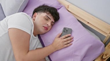 Genç adam elinde akıllı telefonuyla yatak odasında uyuyor. Modern gündelik hayata bir göz atıyor..