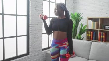 Afrikalı bir kadın evde dambıllarıyla egzersiz yapıyor. Parlak bir oturma odası, kanepesi ve modern bir dekorasyonu var..