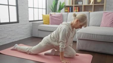 Olgun sarışın bir kadın yoga yapıyor parlak, rahat bir oturma odasında, koltuklu pembe bir minderde poz veriyor.