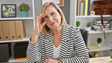 Modern bir ofiste oturan çizgili ceketli, gülümseyen olgun bir kadın..