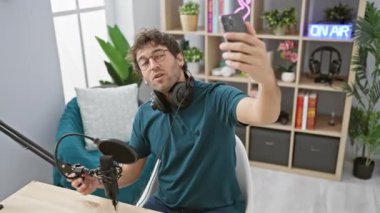 Podcast stüdyosunda kulaklıklı bir adam canlı, modern yayın ortamını gösteren bir selfie çekiyor..
