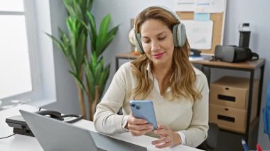 Genç İspanyol kadın ofiste akıllı telefon kullanırken kulaklıkla müzik dinlemekten hoşlanıyor.