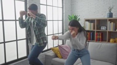 Bir adam ve bir kadın, beyaz bir kanepe ve kitaplıkla, parlak bir oturma odasında şakalaşarak boks yapıyorlar..