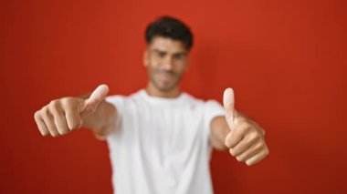 Sakallı neşeli genç bir adam kırmızı duvara başparmak işareti yapıyor.