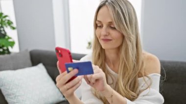 Akıllı telefonunu ve kredi kartını kullanarak rahat bir oturma odasında online alışveriş yapan genç bir kadın..