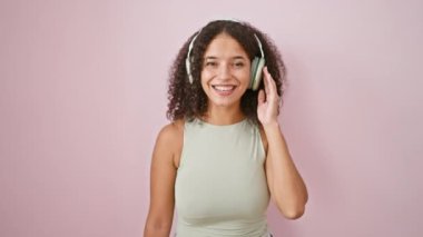 Genç, güzel, İspanyol bir kadın müzik dinliyor. İzole edilmiş pembe arka planda başparmak hareketi yapıyor.