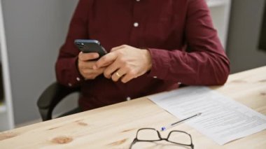 Ofisteki İspanyol adam akıllı telefon kullanıyor, günlük kıyafetleri ve odaklanmış ifadesiyle, yoğun bir profesyonel hayatı somutlaştırıyor..