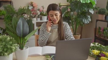Genç bir İspanyol kadın, bitkilerle dolu bir evde birden fazla iş yapıyor. Kırmızı bir telefonla konuşurken dizüstü bilgisayarla çalışıyor..