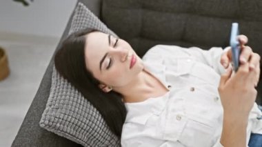 Genç bir İspanyol kadın, bir dairede rahatça uzanır. Oturma odasında akıllı telefon kullanır. Güzellik ve rahatlama yayar..