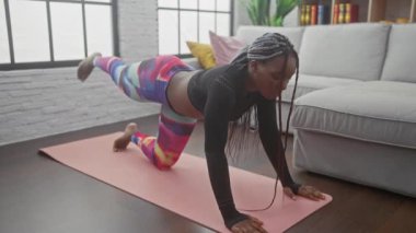 Afrika kökenli Amerikalı bir kadın oturma odasında egzersiz yapıyor. Kapalı bir yerde yoga yapıyor. Renkli tayt ve siyah bluz giyiyor..