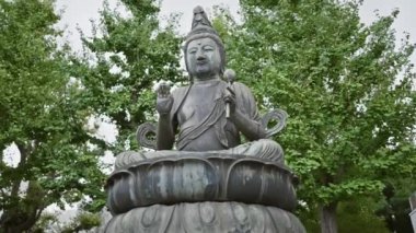 Budist bir ilahın bronz heykeli yemyeşil ağaçlarla çevrili sakin bir Japon parkında huzur içinde duruyor..