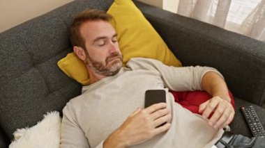 Orta yaşlı, sakallı bir adam rahat bir oturma odasında akıllı telefon kullanıyor..