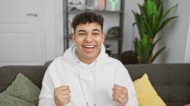 A cheerful young man with curly hair in a casual hoodie celebrates an achievement in a cozy modern living room. clipart