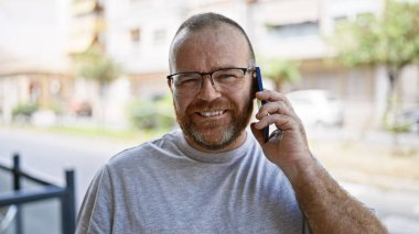 Yakışıklı, sakallı, beyaz bir adam akıllı telefonuyla kendinden emin bir şekilde sohbet ediyor, eğlenceli ve güneşli şehir sokaklarına dalmış, sıcacık bir gülümsemeyle sıradan, serin bir hayat tarzını canlandırıyor.