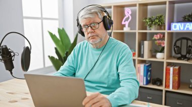 Kulaklık takan olgun bir adam, parlak bir podcast stüdyosunda mikrofon ve dizüstü bilgisayarla bir masada oturuyor..