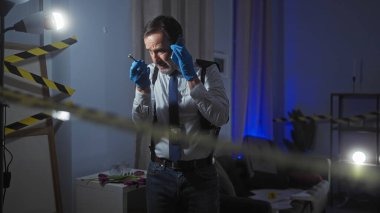 Olgun takım elbiseli bir adam telefon ve kalem tutuyor, loş, dikkat bantlı bir suç mahallinde ipuçlarını analiz ediyor..