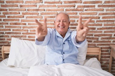 Orta yaşlı, kır saçlı, zafer jesti yapan bir adam yatak odasında oturuyor.
