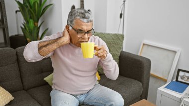 Orta yaşlı, boynu ağrıyan, kanepede kahve içen bir adam.