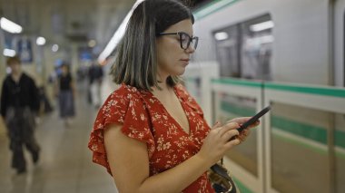 Göz kamaştırıcı İspanyol kadın, gözlüklü, yeraltı tren istasyonunda duruyor, metro yolculuğunu tahmin ediyor, telefonuna dalmış.