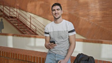 Akıllı telefonlu yakışıklı genç adam şehir sokaklarının merdivenlerinde dikilmiş gülümsüyor..