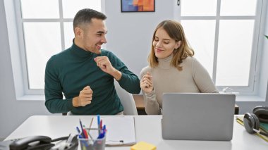 A smiling man and woman fist bump in a bright office, symbolizing teamwork, with a laptop and stationery on the desk. clipart