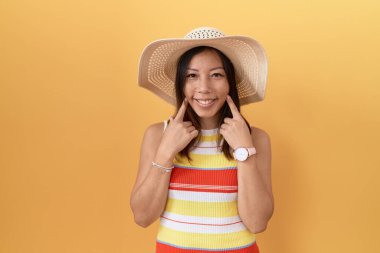 Sarı arka planda sarı şapkalı orta yaşlı Çinli kadın ağzı açık gülümsüyor, parmakları işaret ediyor ve neşeli bir gülümseme sergiliyor. 