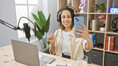 A smiling young woman with headphones takes a selfie in a modern radio studio setup, showcasing media technology. clipart