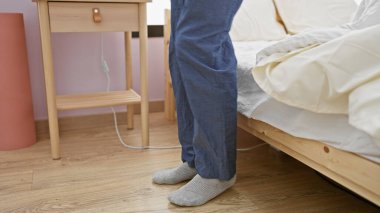 Bir adam rahat bir yatak odasında, pijamalarının yanında duruyor. Bu da evde rahat bir sabah rutini olduğunu gösteriyor..