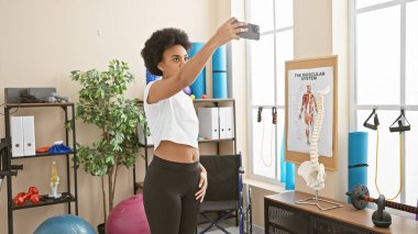 Özgüvenli bir Afrikalı Amerikalı kadın spor salonunda özçekim yapıyor. Arka planda egzersiz ekipmanları var..