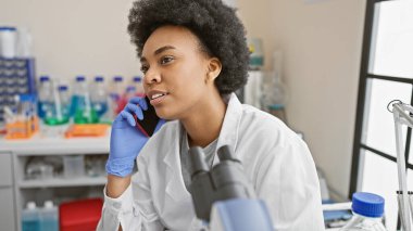 Afrika kökenli Amerikalı kadın bilim adamı telefonda konuşurken iyi donanımlı bir laboratuvarda çalışıyor.