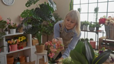 Sarışın bir kadın canlı bir çiçekçinin içinde çiçeklerin fotoğraflarını çekiyor, yemyeşil çiçeklerle çevrili..
